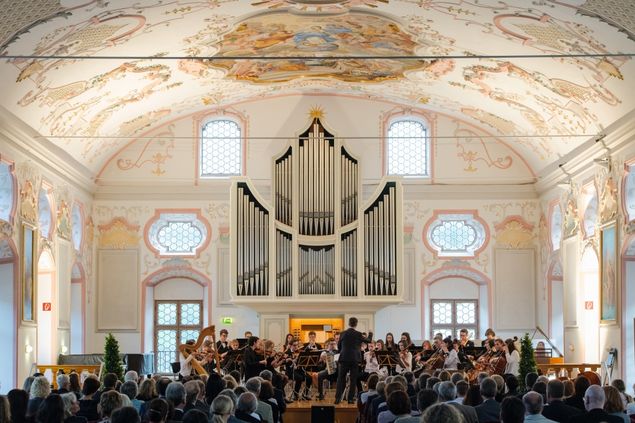Publikum und Orchester in der barock geschmückten Aula des Kurfürst-Maximilian-Gymnasiums Burghausen mit Orgel im Hintergrund